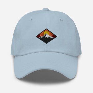 May include: A light blue baseball cap with an embroidered patch of a mountain range with a setting sun. The patch is in a diamond shape with black outlines and red, orange, and yellow colors.