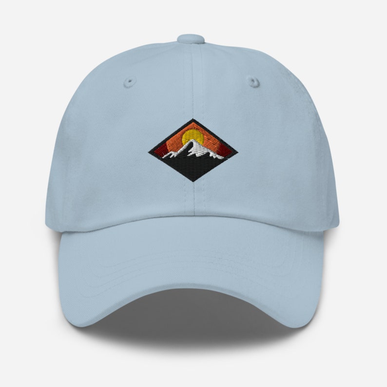 May include: A light blue baseball cap with an embroidered patch of a mountain range with a setting sun. The patch is in a diamond shape with black outlines and red, orange, and yellow colors.