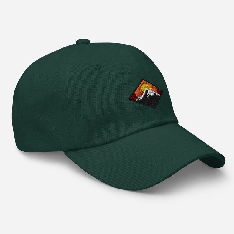 May include: A dark green baseball cap with a diamond-shaped patch featuring a mountain range and a setting sun. The patch is embroidered with orange, yellow, red, and white thread.
