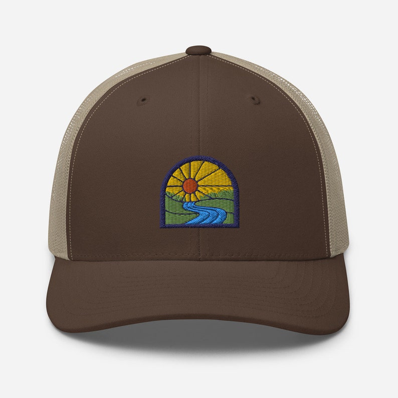 May include: A brown and beige trucker hat with a patch of a sun setting over a river and hills. The patch is in a stained glass style.