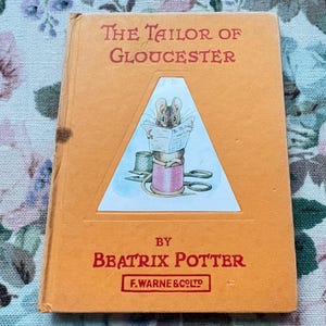 Puede incluir: Una copia vintage de "The Tailor of Gloucester" de Beatrix Potter. El libro tiene una portada naranja con el título en rojo y una ilustración triangular de un ratón leyendo un periódico. También son visibles el nombre del autor y el editor.