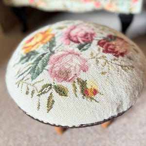 May include: A small, round footstool with a floral tapestry design. The top features a cream-coloured background with embroidered roses in shades of pink, yellow, and red, surrounded by green leaves. The stool has a dark brown trim and wooden legs.