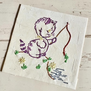 Puede incluir: Cuadrado bordado con un gato morado pescando con una línea marrón. El gato tiene un lazo amarillo y está rodeado de hierba verde y flores amarillas. El fondo es de tela blanca.