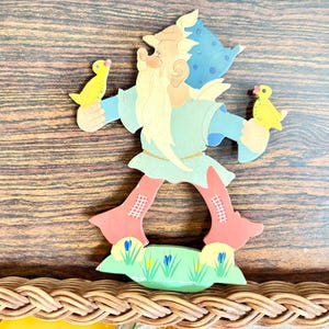 May include: A wooden cutout of a whimsical gnome figure holding two yellow ducklings. The gnome wears a blue hat with white polka dots, a light blue tunic, and red trousers. The base features green grass and blue flowers.