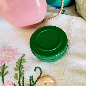 Puede incluir: Un pequeño recipiente redondo de plástico verde oscuro con tapa plana. El recipiente está colocado sobre una tela blanca con flores bordadas en rosa y morado con tallos y hojas verdes, cerca de un objeto rosa y un botón de concha.