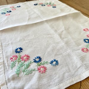 Può includere: Runner da tavolo in lino bianco con ricamo floreale. Il ricamo presenta fiori rosa e blu con steli verdi. Il runner ha un bordo cucito e viene esposto su una superficie di legno.