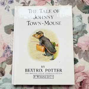 Peut inclure: Un livre intitulé "The Tale of Johnny Town-Mouse" de Beatrix Potter. La couverture présente une illustration d'une souris en manteau et chapeau, portant une valise. Le livre est posé sur un fond floral.