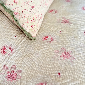 Pode incluir: Uma colcha acolchoada de cor creme com um padrão repetitivo de pequenos motivos florais vermelhos. Uma almofada a condizer com uma borda verde com folhos e uma estampa floral e paisley vermelha repousa sobre o tecido acolchoado, exibindo costuras.