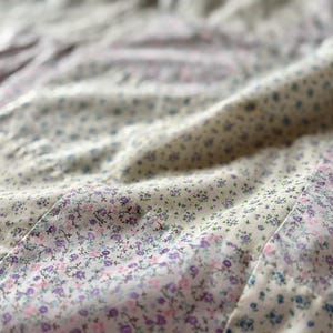 May include: Close-up of a floral fabric with a cream background. The fabric is covered in small, delicate flowers in shades of purple, pink, and blue. The fabric is slightly wrinkled, suggesting it is part of a garment or bedding.