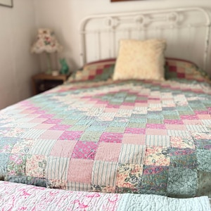 Puede incluir: Una colcha de patchwork con un patrón de diamantes compuesto por pequeños cuadrados en varios tonos de rosa, verde salvia y blanco, con estampados florales, de rayas y de cuadros vichy. La colcha cubre una cama con cabecero de metal blanco.