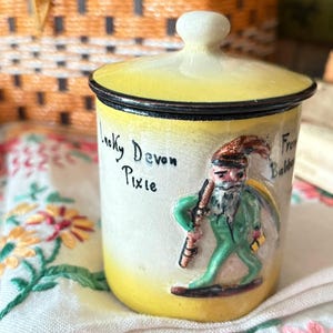 May include: A yellow and white ceramic jar with a matching lid, featuring a raised relief figure of a pixie in a green outfit and a brown hat. The text "Lucky Devon Pixie" and "From Babbacombe" is hand-painted in black on the front of the jar.