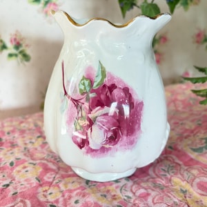 May include: A white ceramic vase with a ruffled top and a floral design. The vase features a large pink rose with green leaves. The rim is trimmed with gold. A decorative piece for the home.
