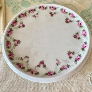 May include: A white, circular ceramic plate featuring a floral pattern. Pink roses and green leaves decorate the edge of the plate. The plate is displayed on a light-coloured surface.