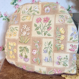 Può includere: Una teiera floreale con un design patchwork. La teiera presenta fiori ricamati in rosa, giallo e viola su uno sfondo beige. La teiera misura circa 30 cm di larghezza e 25 cm di altezza.
