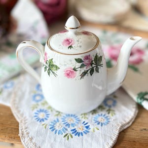 May include: A white porcelain teapot with a gold trim and handle, decorated with pink roses and green foliage. The teapot is placed on a white doily with blue floral embroidery. A charming piece for tea time.