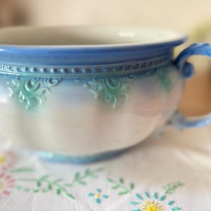 May include: A ceramic chamber pot featuring a white base with a light blue rim and handle. The exterior is decorated with embossed scrollwork patterns in a soft teal colour, accented by a beaded texture along the upper edge of the rounded vessel.