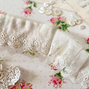 Peut inclure: Une longueur de dentelle blanc cassé présentant un motif floral délicat brodé le long du bord festonné. Le tissu est une maille fine et semi-transparente avec une texture froncée, mettant en valeur des détails de broderie complexes.