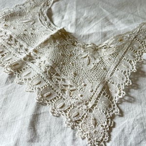 Puede incluir: Cuello de encaje crema antiguo con un delicado diseño calado. El cuello presenta intrincados patrones florales y de hojas a lo largo de los bordes y un ribete festoneado. El encaje está colocado sobre una superficie de lino blanco, mostrando sus finos detalles.