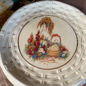 May include: Cream-coloured decorative plate with a woven basket-like rim. The centre features a garden scene with flowers, a path, and an archway. The plate has a gold-coloured border.