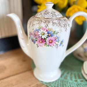 May include: White ceramic coffee pot with a floral design featuring pink, blue, and white daisies. The lid and rim have gold accents. A matching teacup and saucer set is visible in the background.