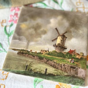 Può includere: Una piastrella quadrata in ceramica con un paesaggio dipinto raffigurante un mulino a vento situato vicino a uno specchio d'acqua con un molo in legno. La scena include rive erbose verdi, piccoli edifici con tetti rossi e un cielo nuvoloso.