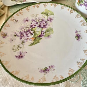 Könnte beinhalten: Ein weißer Keramikteller mit grünem Rand und floralem Design. Der Teller zeigt eine zentrale Ansammlung von lila Blumen mit grünen Blättern und kleinere florale Akzente am Rand. Dekoratives Geschirr.