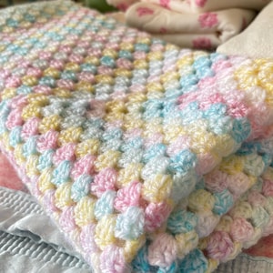 Könnte beinhalten: Eine gehäkelte Decke in Pastellfarben, Rosa, Gelb und Blau. Die Decke ist gefaltet und zeigt das strukturierte Stichmuster. Die sanften Farben und die handgefertigte Qualität deuten auf eine Babydecke oder einen dekorativen Überwurf hin.