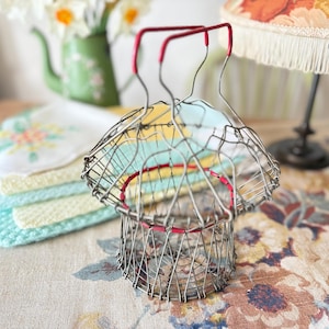 May include: A retro wire egg basket with red detailing. The basket is constructed from silver-coloured wire and has a rounded form. The handle is also red. The background features a green teapot, a floral lampshade, and pastel-coloured cloths.