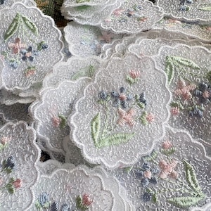 May include: A collection of white lace appliques with a scalloped edge. Each piece is decorated with embroidered flowers in pink, blue, and green. The floral embroidery adds a delicate touch to the lace.