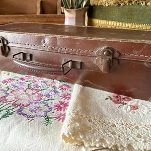 May include: A brown leather suitcase with metal clasps and a handle. The rectangular case shows signs of age. It sits on a white embroidered cloth with floral patterns. The image evokes a sense of classic, antique travel.