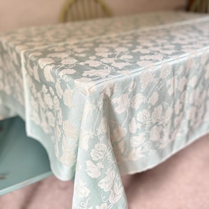 May include: A rectangular tablecloth in a soft sage green color featuring a woven, tonal floral pattern in a lighter cream shade. The fabric drapes over the edges of a table, displaying a repeating botanical design of flowers and leaves.