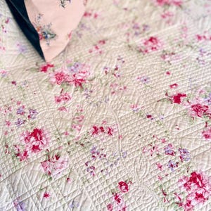 May include: A cream-colored quilted bedspread featuring a repeating floral pattern with vibrant pink roses, small purple blossoms, and green foliage. The fabric has a textured grid-like stitching pattern throughout, with a corner of a matching pillow.