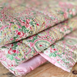 May include: A folded quilt featuring a vibrant floral and paisley pattern in shades of pink, green, and yellow. The fabric has a detailed botanical print with intricate swirls and blossoms, finished with a smooth, solid light pink satin border.