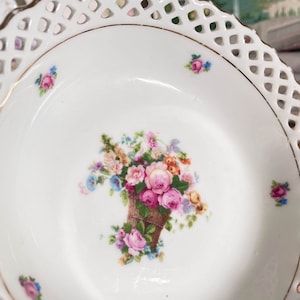 May include: A white porcelain bowl with a delicate lace-like rim and a floral design featuring a basket of pink, yellow, and blue flowers. The bowl has a gold trim around the edge.