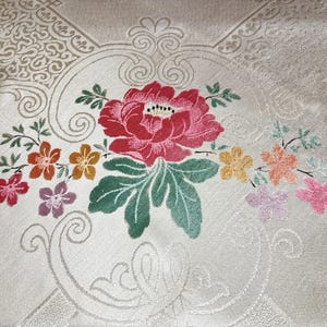 May include: Close-up of a cream fabric featuring an embroidered floral design. A large red peony is the focal point, surrounded by smaller flowers in shades of orange, yellow, and pink. Green leaves and stems complete the floral arrangement.
