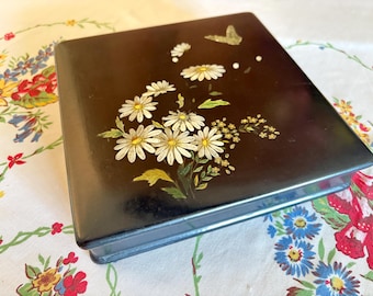 Può includere: Una scatola quadrata nera con coperchio incernierato, decorata con margherite bianche, accenti gialli e una farfalla dorata. Il motivo floreale è dipinto sulla superficie superiore della scatola. La scatola è appoggiata su una tovaglia a motivi floreali.