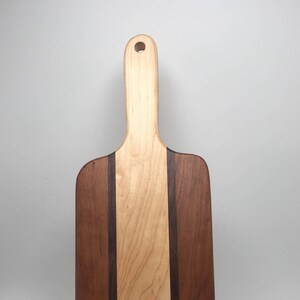 May include: A rectangular wooden cutting board with a handle. The board features a light-colored wood center panel, flanked by darker wood on the sides, and a dark wood border. A hole is drilled in the handle for hanging.