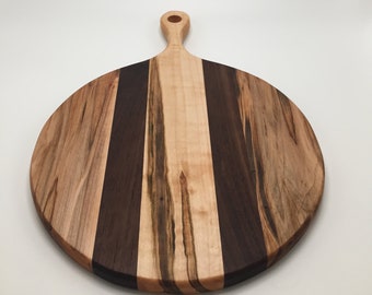 May include: Round wooden cutting board with a handle. The board features alternating vertical stripes of light and dark wood. The handle has a hole for hanging. The board is approximately 12 inches in diameter.