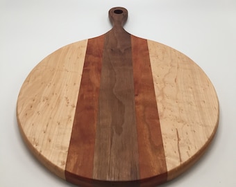 May include: A round wooden cutting board with a handle. The board features a light-colored wood with darker wood stripes in the center. The handle is a darker brown color. The board is approximately 14 inches in diameter.
