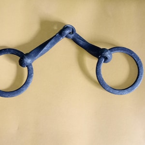 May include: A dark grey metal horse bit with two circular rings connected by angled bars. The bit is made of forged metal and is displayed against a tan background. The rings are approximately 10 cm in diameter.