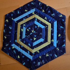 K&ouml;nnte beinhalten: Ein sechseckiger blauer, gesteppter Tischl&auml;ufer mit einem geometrischen Muster aus gelben und blauen Streifen. Der Stoff hat einen Blumendruck mit kleinen wei&szlig;en Bl&uuml;ten und gr&uuml;nen Stielen.