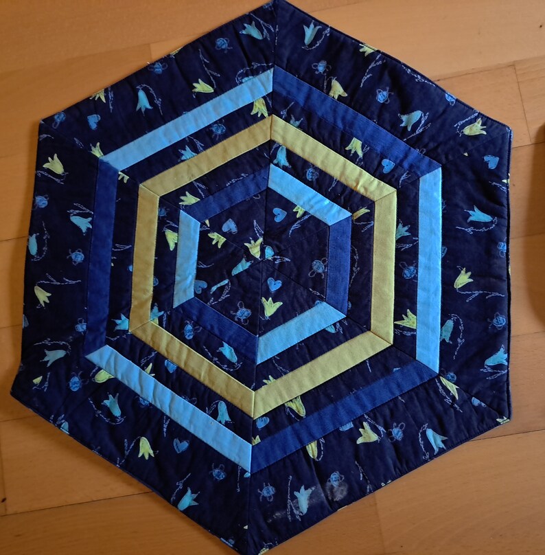 K&ouml;nnte beinhalten: Ein sechseckiger blauer, gesteppter Tischl&auml;ufer mit einem geometrischen Muster aus gelben und blauen Streifen. Der Stoff hat einen Blumendruck mit kleinen wei&szlig;en Bl&uuml;ten und gr&uuml;nen Stielen.