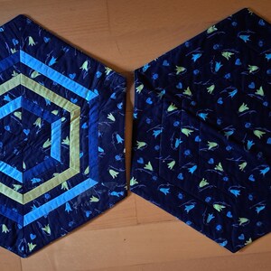 K&ouml;nnte beinhalten: Zwei sechseckige Stoff-Platzsets mit einem blauen und gr&uuml;nen Blumenmuster. Ein Platzset hat ein geometrisches Design in der Mitte mit blauen, gr&uuml;nen und gelben Streifen.