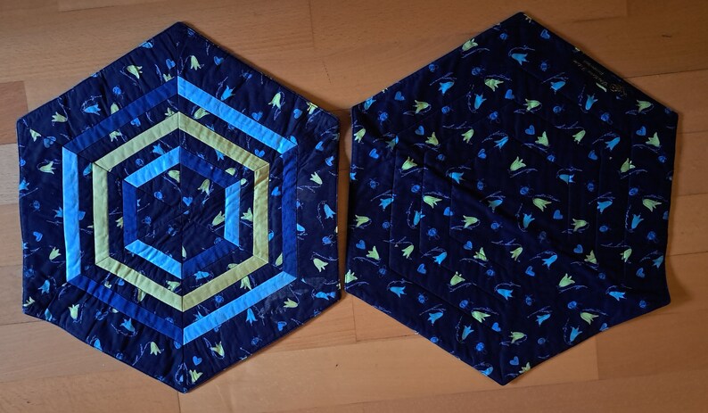 K&ouml;nnte beinhalten: Zwei sechseckige Stoff-Platzsets mit einem blauen und gr&uuml;nen Blumenmuster. Ein Platzset hat ein geometrisches Design in der Mitte mit blauen, gr&uuml;nen und gelben Streifen.