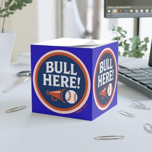May include: A blue sticky note cube featuring the text "BULL HERE!" in white, within a red and orange circle, with a baseball and megaphone graphic. The cube is on a white surface.