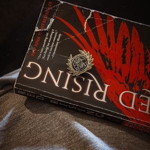 Puede incluir: Un libro negro con texto rojo y blanco que dice "Red Rising" y "Pierce Brown". Un pin dorado con las palabras "Hail Libertas Hail Rexpet" está en el libro.