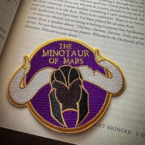 May include: A purple and gold embroidered patch with the text "The Minotaur of Mars". The patch features a stylized image of a Minotaur's head with horns.