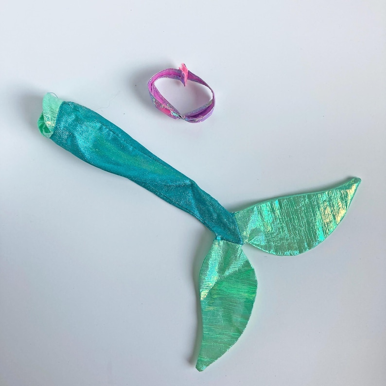 May include: A shiny teal mermaid tail costume piece with a metallic finish and a flared fin, accompanied by a small, circular, pink and purple patterned fabric band. The tail features a smooth texture and a vibrant, reflective green-blue color.