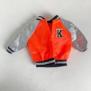 May include: A small, open orange jacket with silver sleeves and a black collar and trim. The jacket has a black and white letter "K" patch on the chest. The jacket is made of a shiny material.