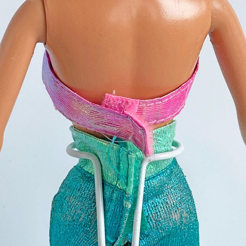 May include: A doll wearing a shiny pink strapless top with a hook-and-loop closure and metallic teal pants with a high waistband. A white wire doll stand supports the figure from the back, encircling the waist over the textured fabric of the clothing.